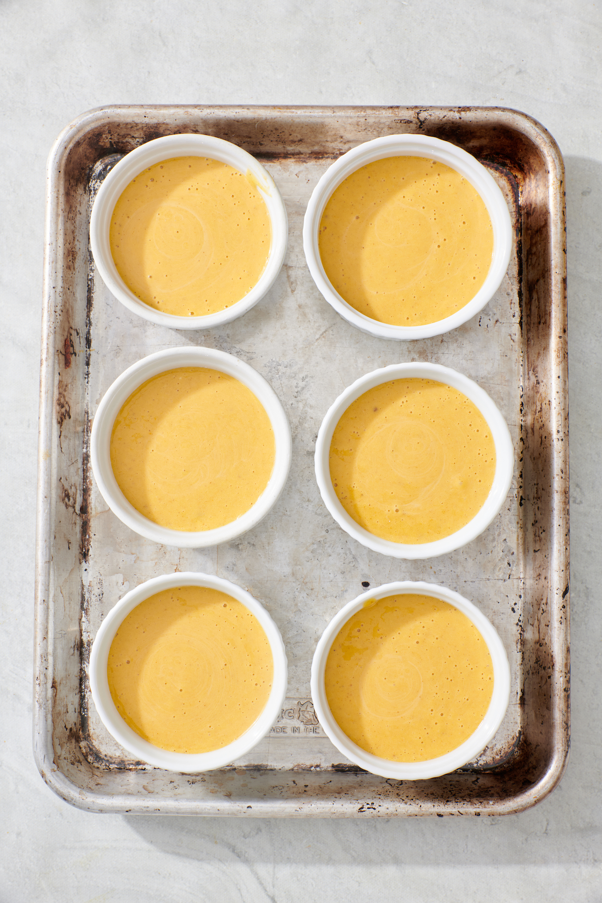 Six ramekins filled with pumpkin cheesecake batter on baking sheet.