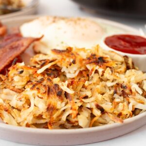 Crispy hash browns on a plate with bacon, eggs, and ketchup.