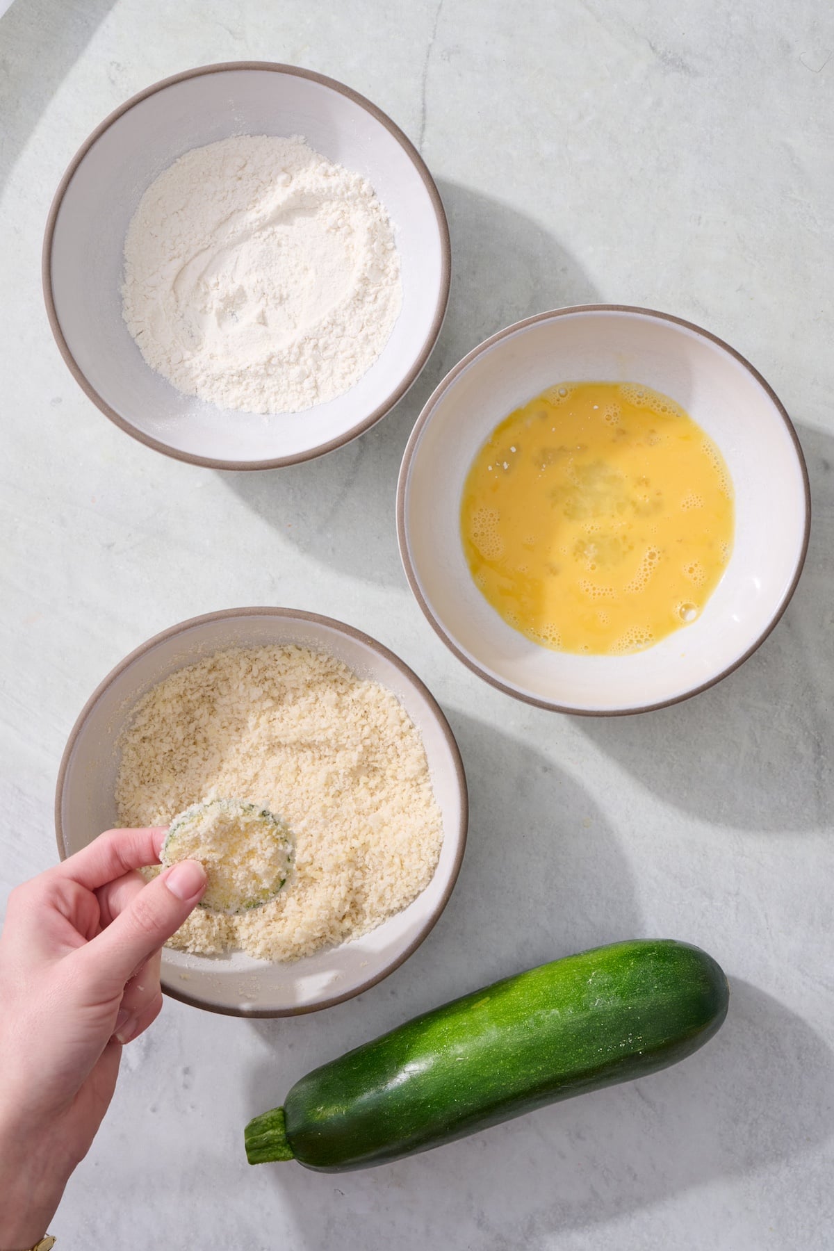 Hand holding a breaded zucchini coin with the breading station bowls.