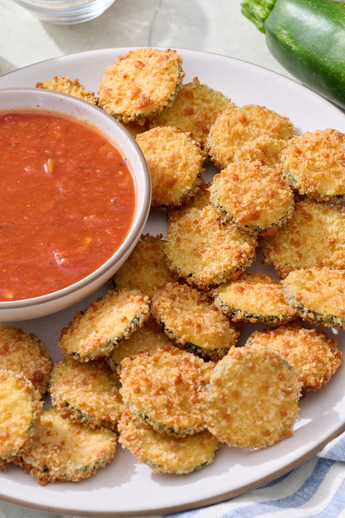 Baked zucchini coins on serving platter with marinara sauce in a bowl on the side.