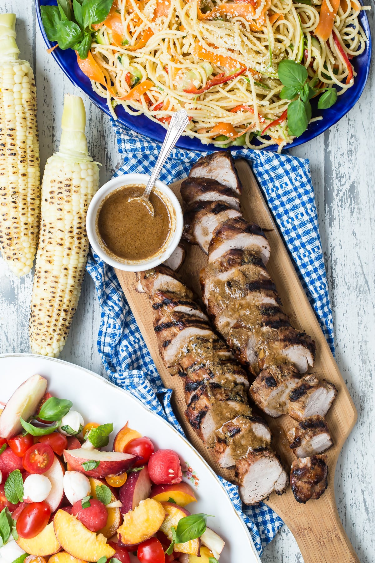 Southern style pork tenderloin on cutting board surrounded by sides of grilled corn, pasta and summer salad.