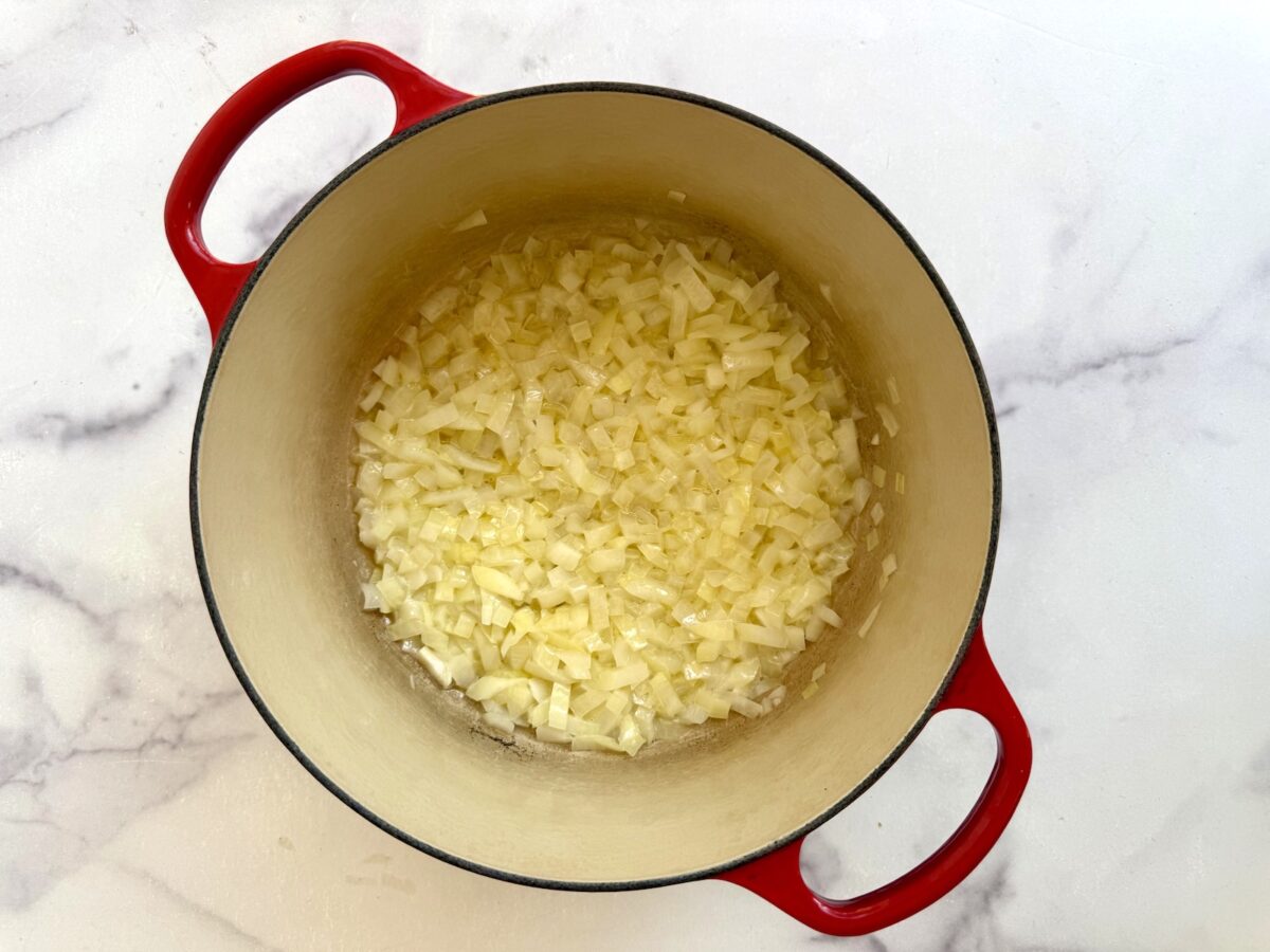 Diced onions cooking in a red enameled cast iron Dutch oven on a white marble surface.