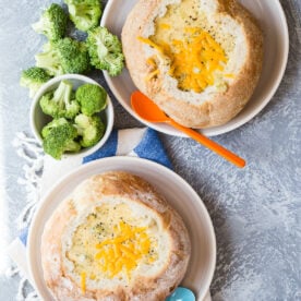 Two bread bowls filled with creamy broccoli cheese soup, served with side bowls of fresh broccoli florets and spoons.