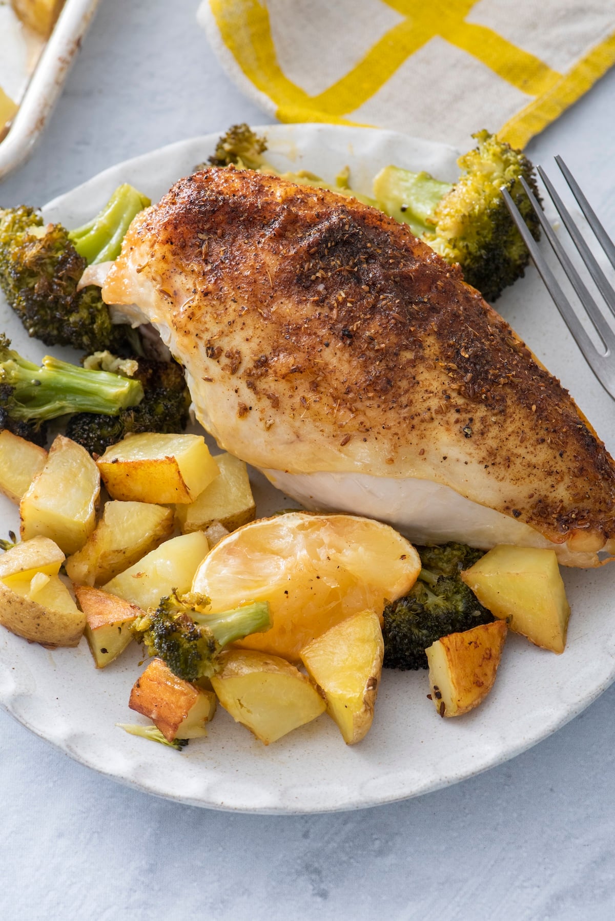A za'atar roasted chicken breast served with crispy potatoes, broccoli, and a roasted lemon half on a plate.