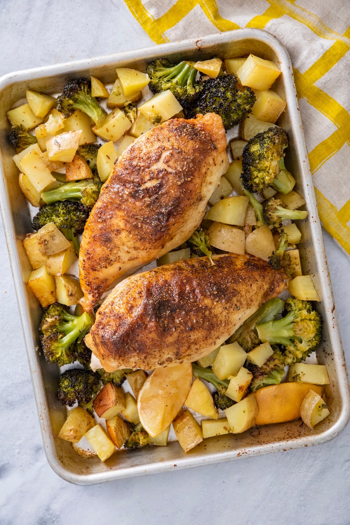 Two za'atar sheet pan chicken breasts roasted with broccoli, potatoes, and lemon on a baking sheet.