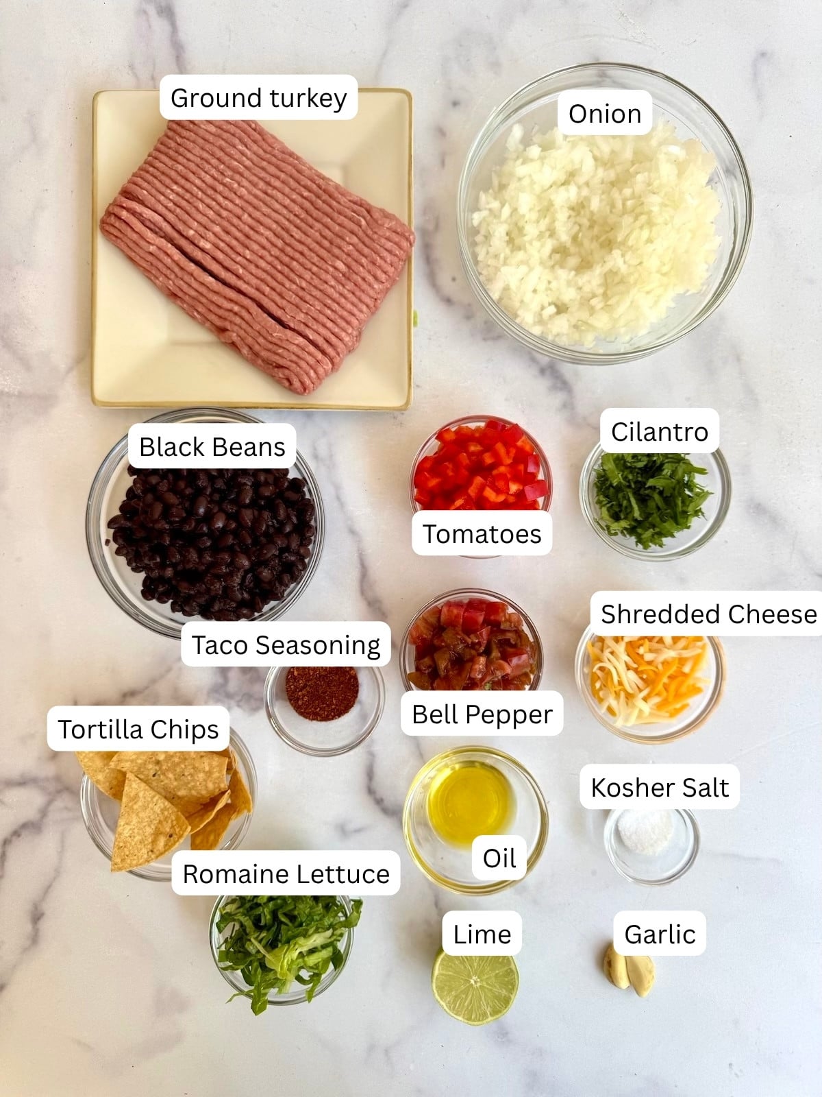 A flat lay of labeled taco bowl ingredients: ground turkey, diced onion, black beans, taco seasoning, tortilla chips, romaine lettuce, tomatoes, oil, bell pepper, lime, garlic, kosher salt, shredded cheese, and cilantro.
