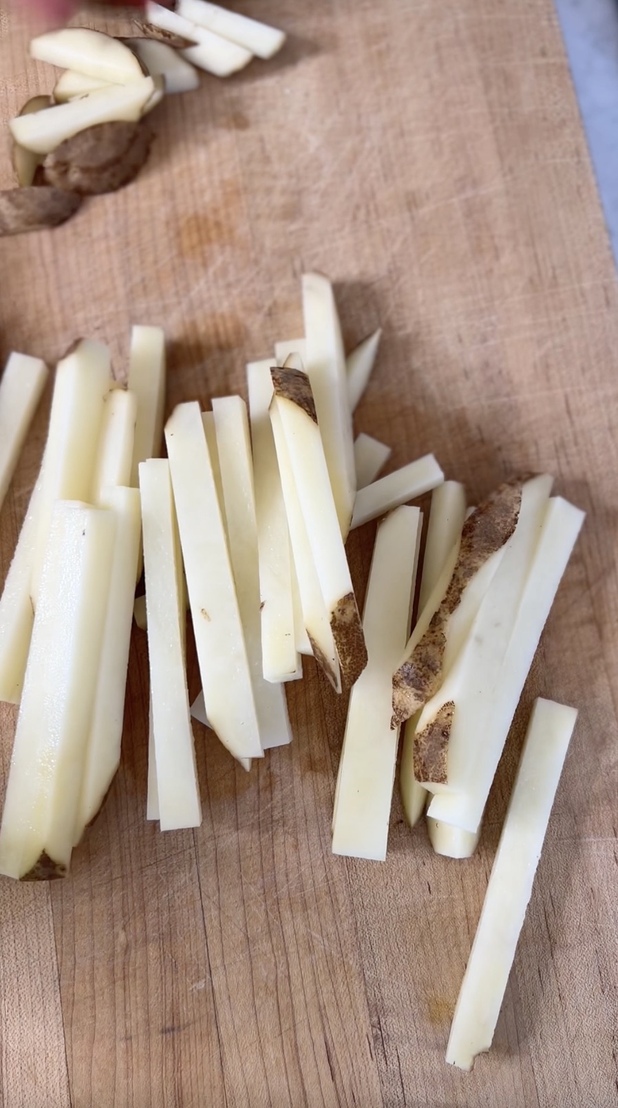 Potatoes cut into fries on wooden cutting board.