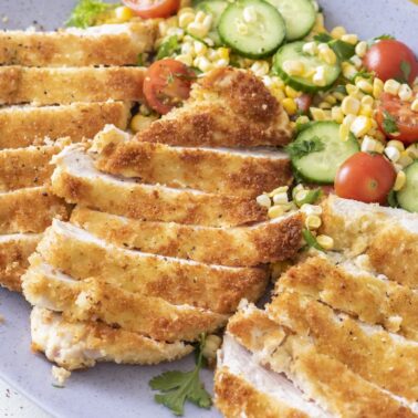 Sliced crispy chicken paillard on a purple platter with a summer salad of corn, cucumber, and cherry tomatoes.