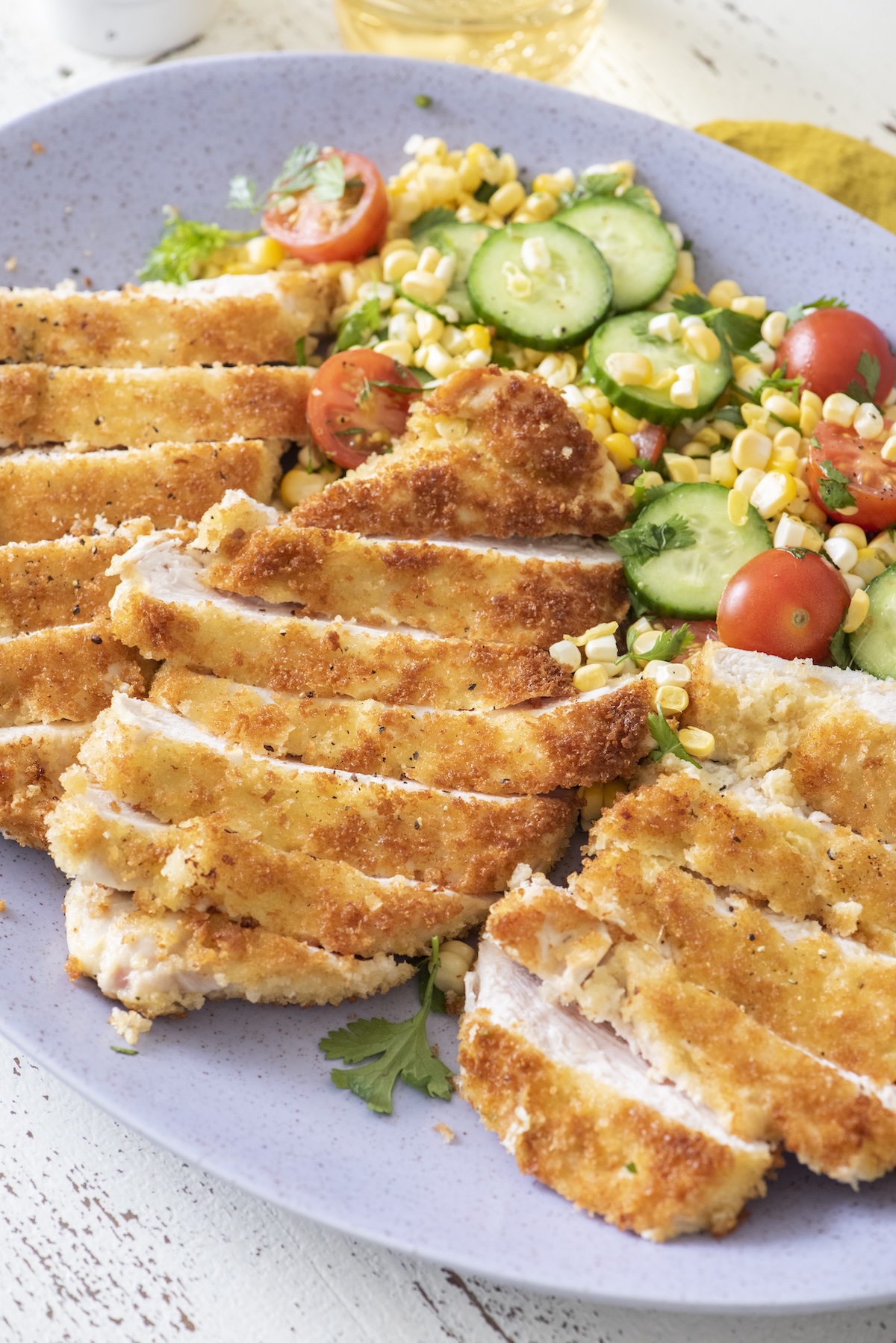 Sliced crispy chicken paillard on a purple platter with a summer salad of corn, cucumber, and cherry tomatoes.