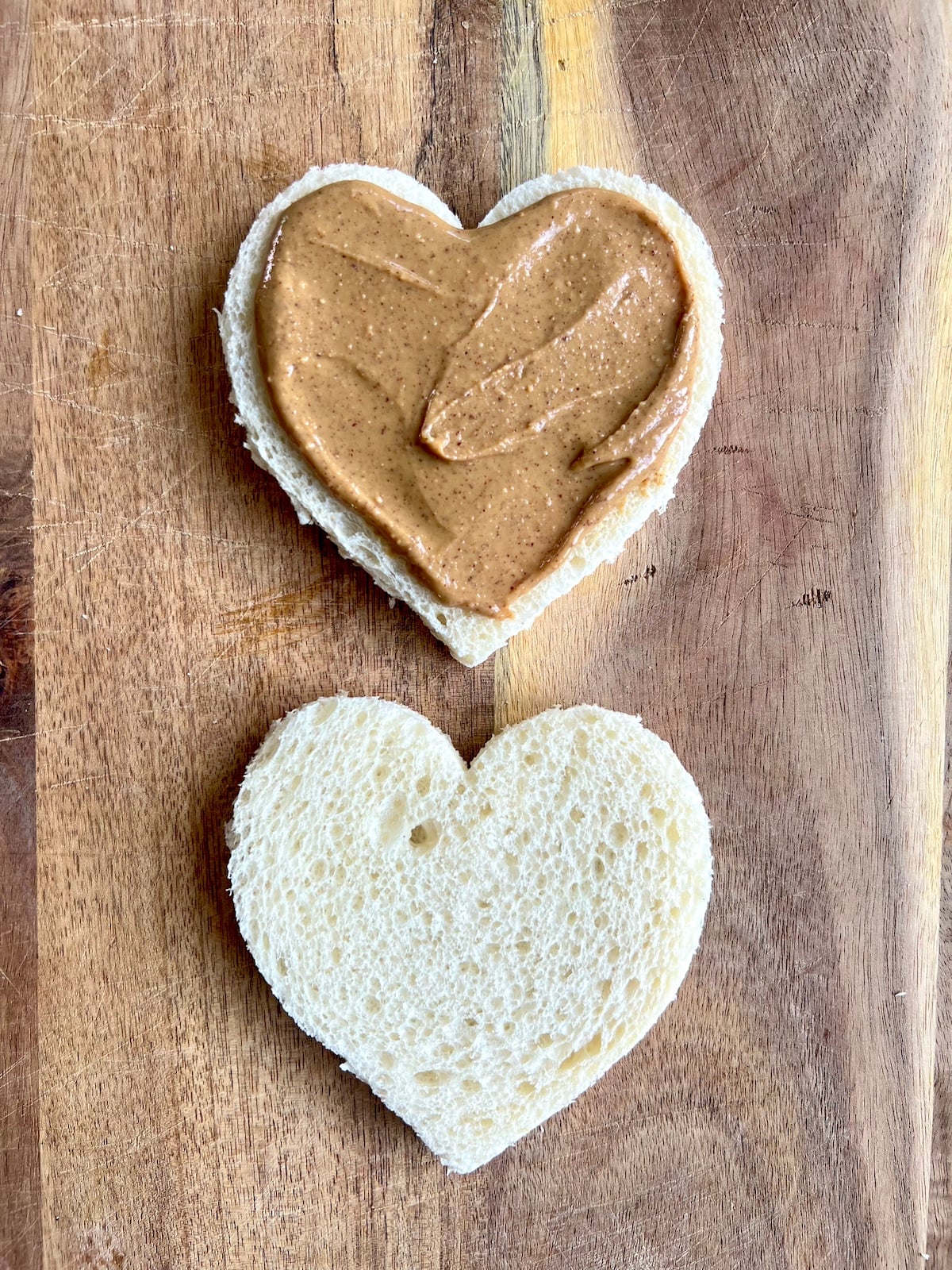 Two pieces of heart shaped bread with peanut butter spread on one.