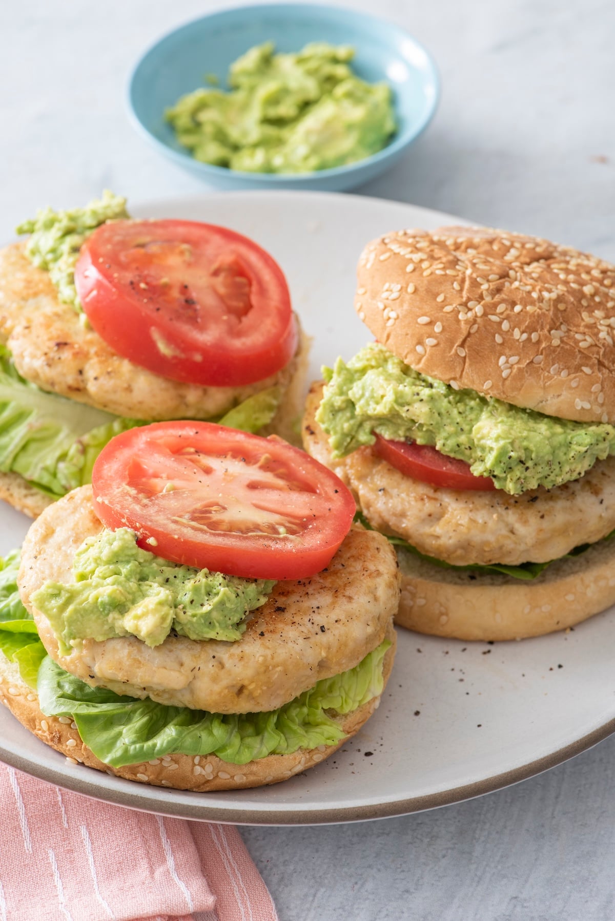 Three juicy turkey burgers with oats on sesame buns topped with guacamole, tomato, and lettuce, served on a white plate with a bowl of guacamole in the background.