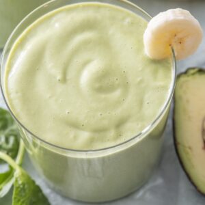 Close-up of a green breakfast smoothie in a glass with a banana slice on the rim, fresh spinach and avocado visible alongside.
