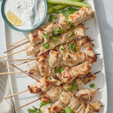 Chicken Tikka skewers on a white platter, served with a cucumber mint raita.