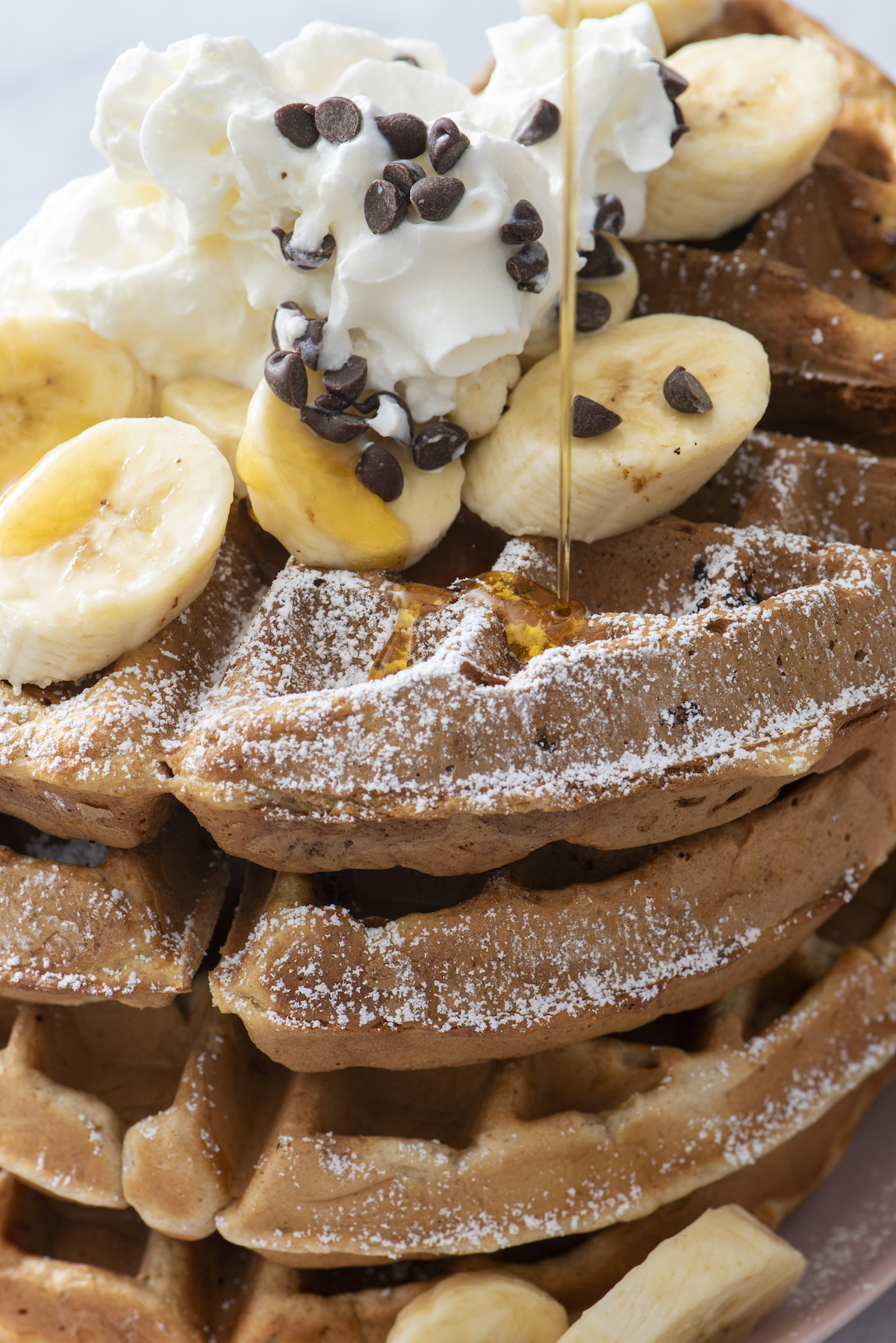 close up of syrup pouring over stack of waffles topped with bananas, chocolate chips and whipped cream