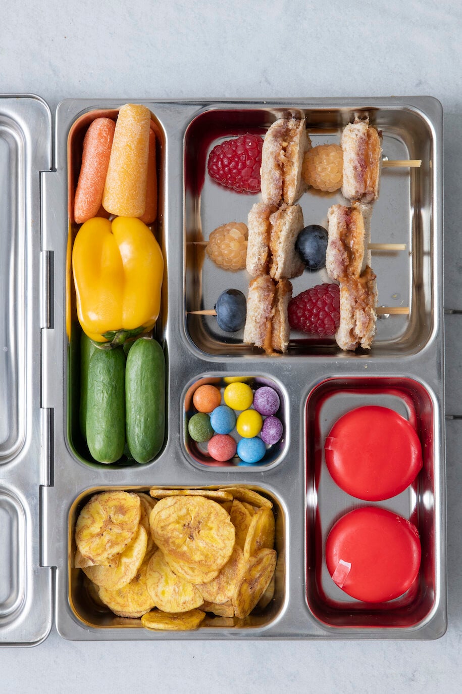 bento style lunch box with peanut butter and jelly skewers, mini cheese wheels, plantain chips, mini cucumbers, mini yellow sweet pepper, carrots and dye free chocolate candies in the center