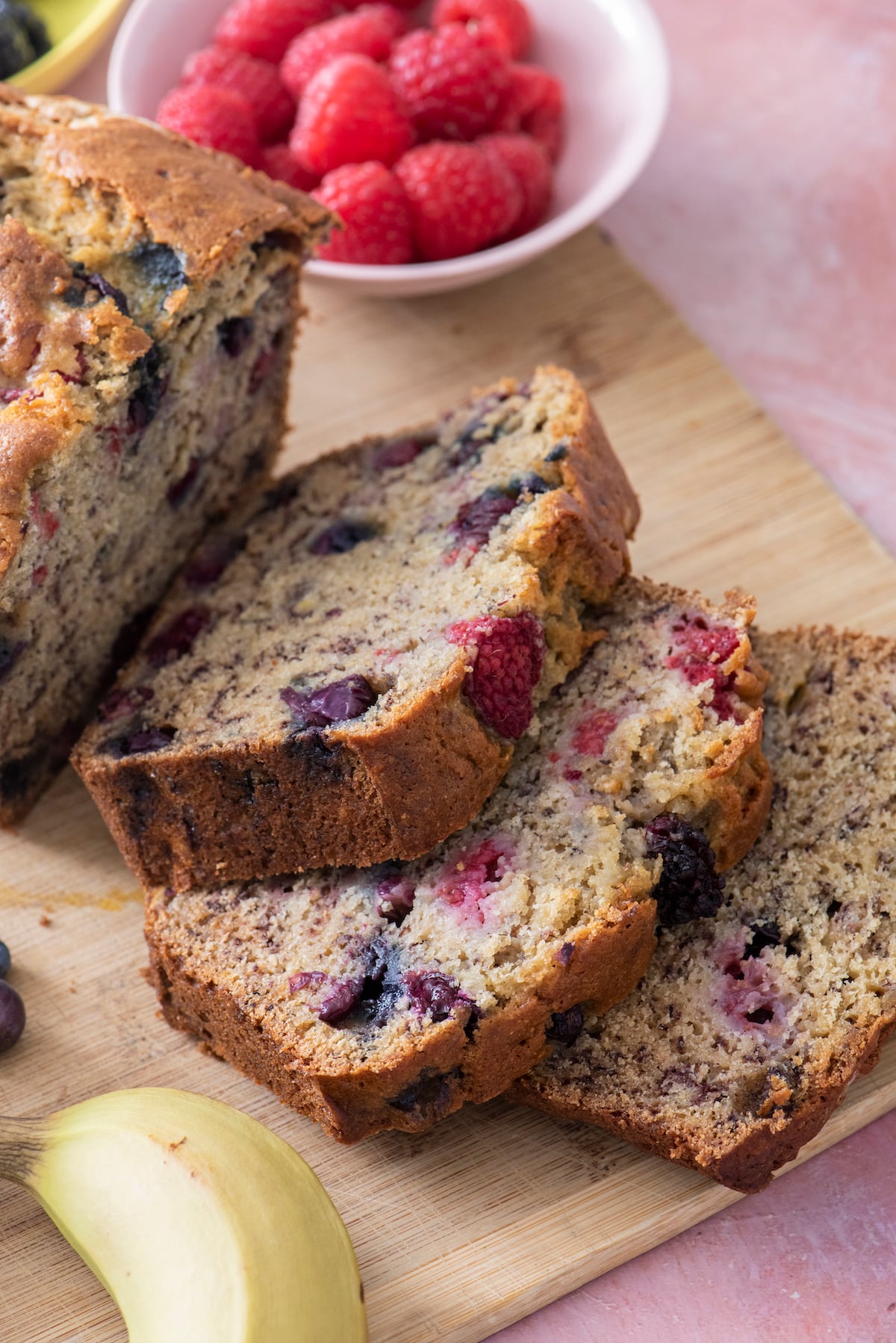 Banana berry bread sliced on wooden cutting board with a banana in the foreground and raspberries in the background.