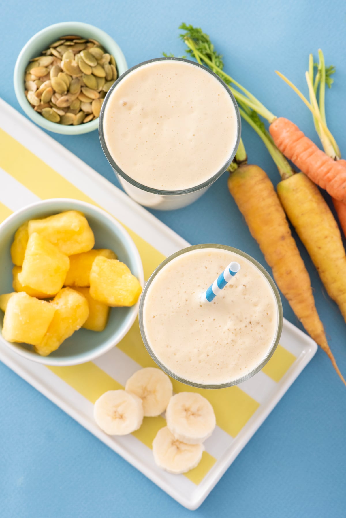 Overhead photo of two smoothies with pumpkin seeds, carrots and pineapple chunks surrounding them.