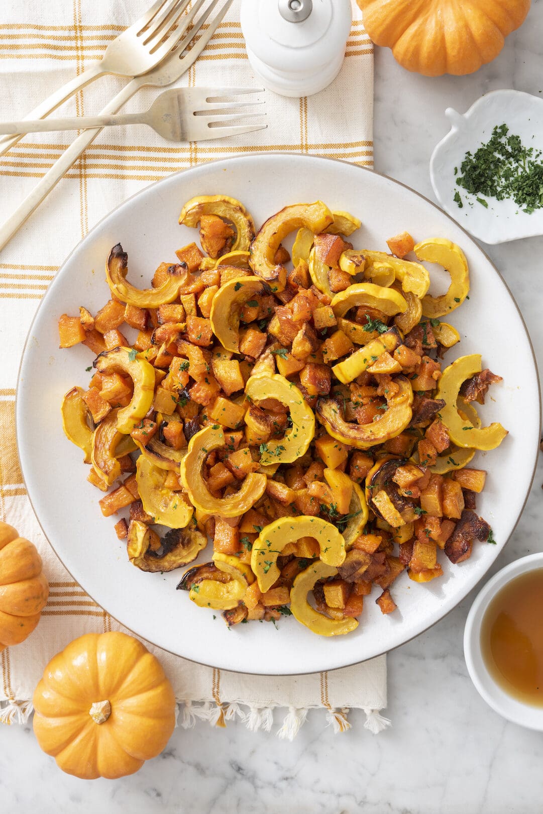 A white plate with roasted delicata squash and sweet potatoes, garnished with herbs. The plate sits on a plaid napkin beside small pumpkins, cutlery, a pepper mill, and a cup of tea.