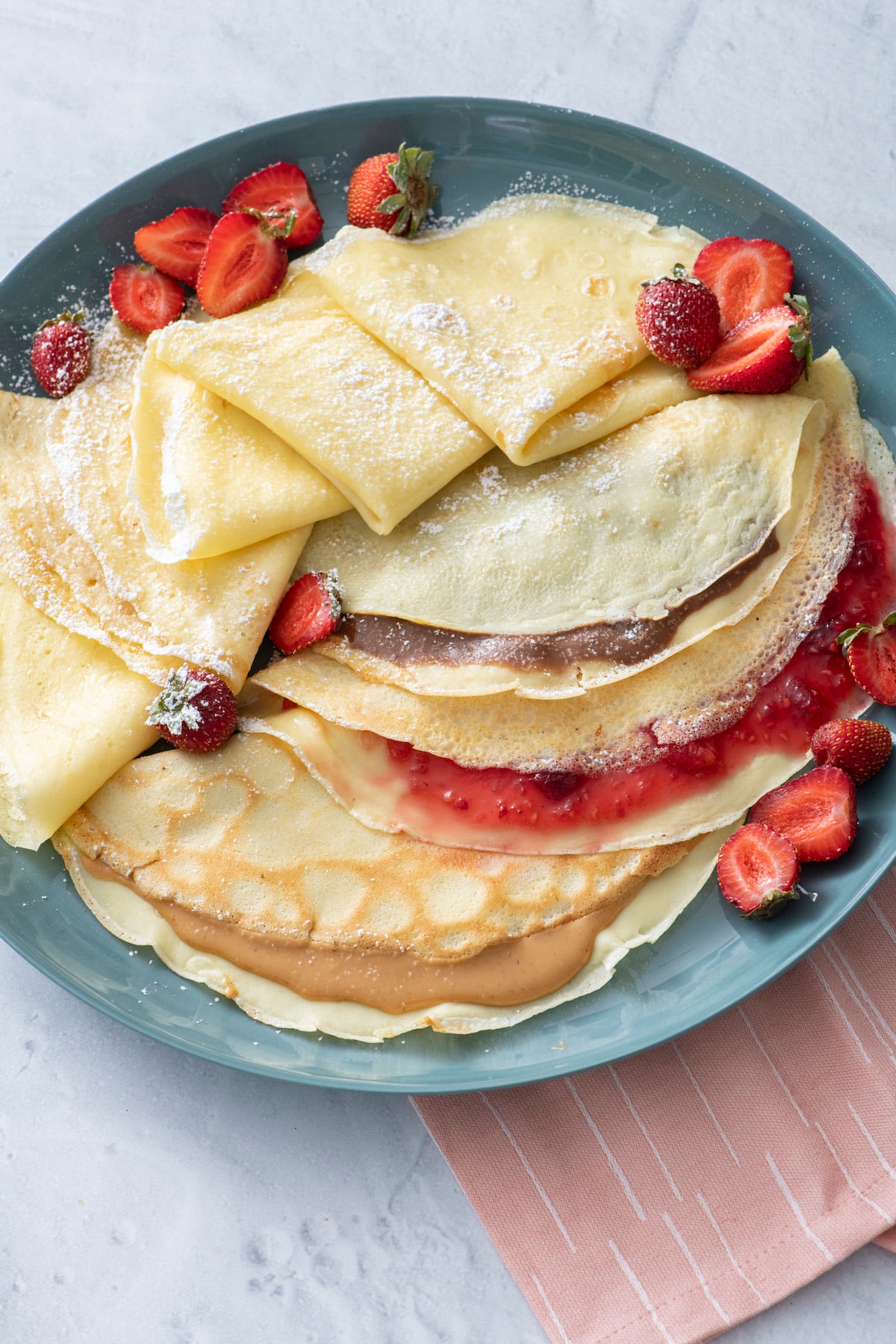 Crepes on blue plate. One filled with Nutella, one with fruit preserves and one with peanut butter.