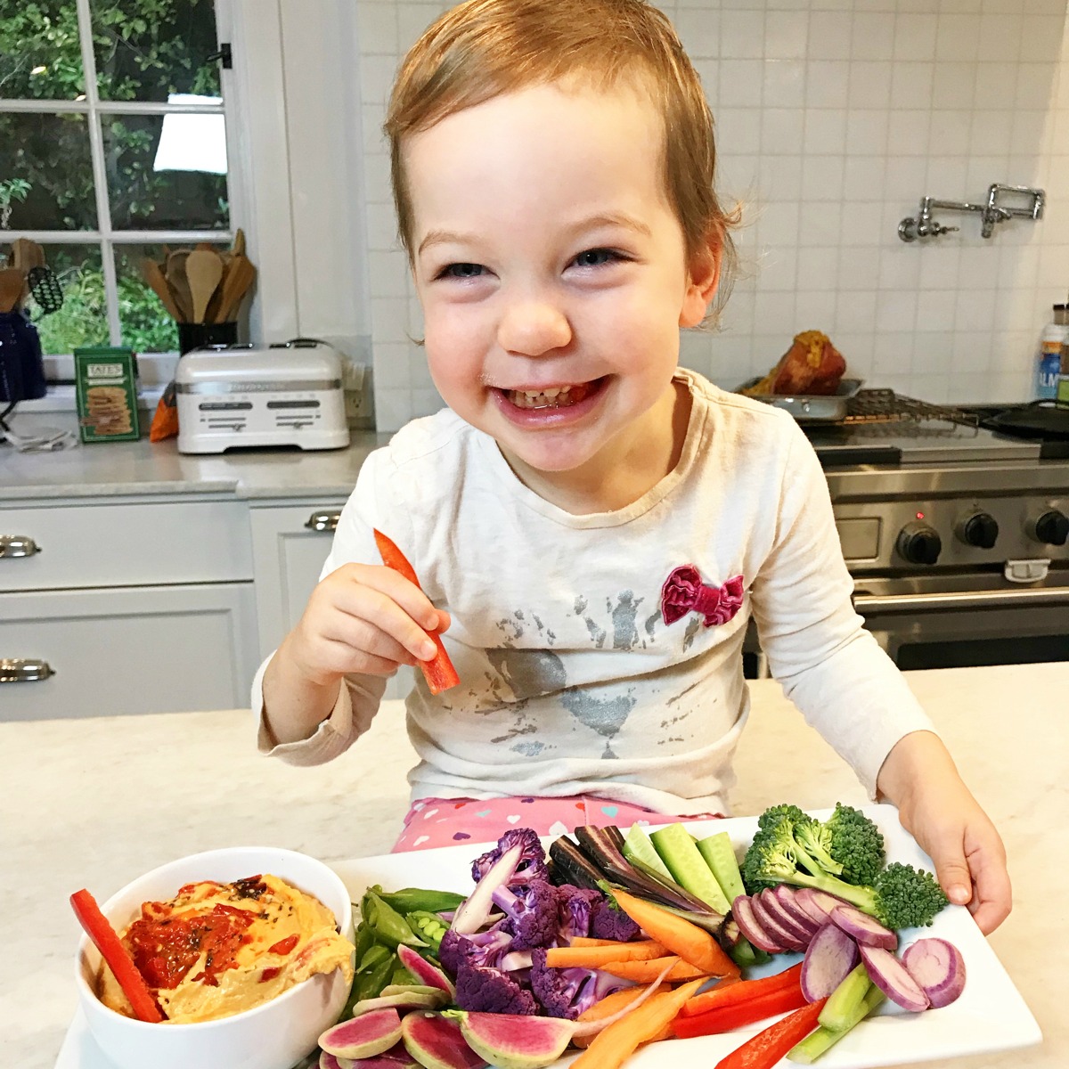 Toddler eating red bell pepper with plate of vegetables in front of her.