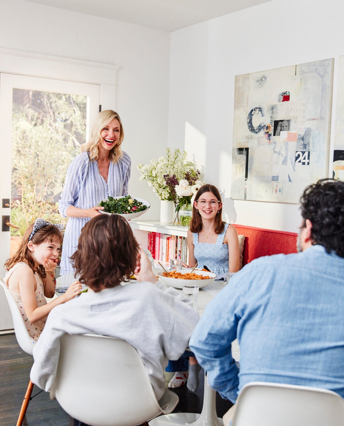 A woman stands serving salad to four people seated around a dining table, smiling and chatting in a bright, modern room with artwork on the wall and sunlight streaming in.
