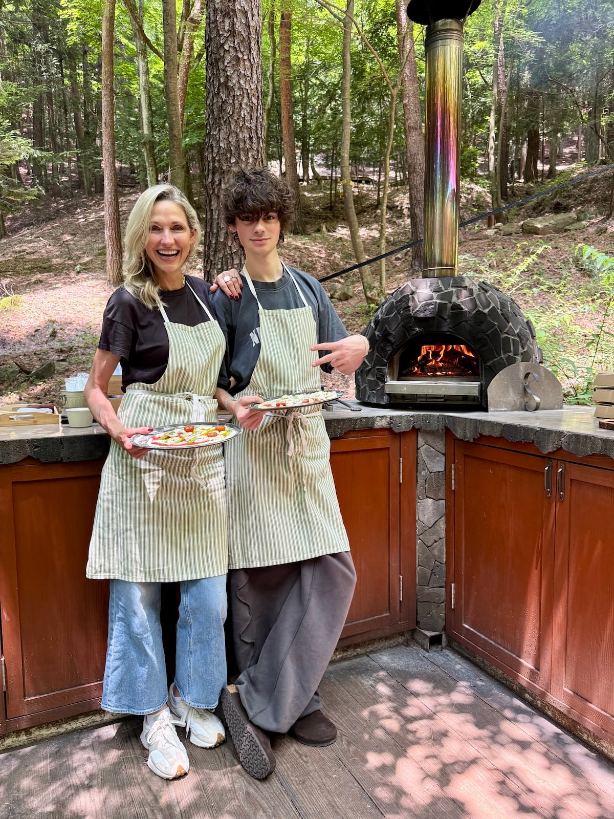 Woman and boy posing with pizzas next to wood fire pizza oven.