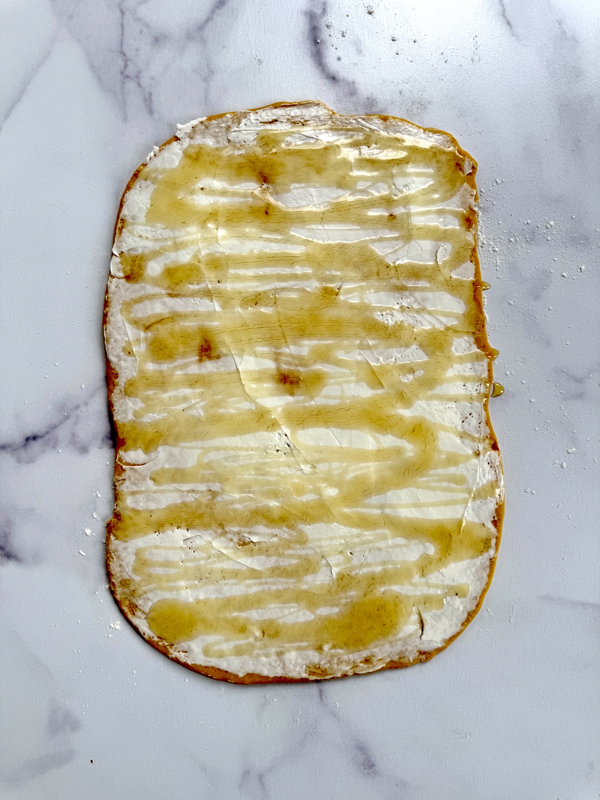 Pumpkin cinnamon roll dough rolled out on a surface. The dough is topped with whipped cream cheese, honey and cinnamon.