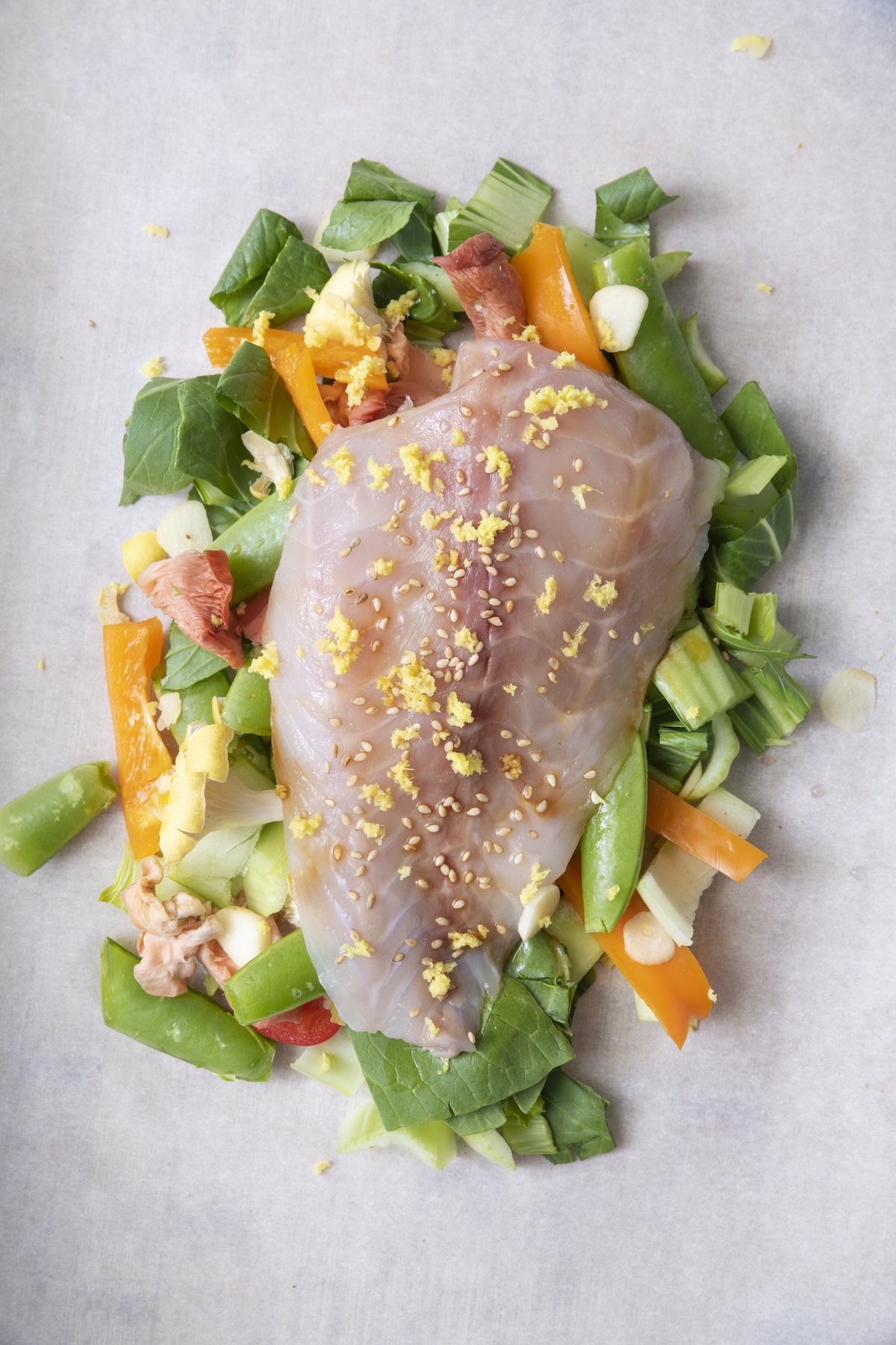 Raw whitefish fillet placed on top of chopped bok choy, snap peas, bell pepper, and shiitake mushrooms on parchment paper before sealing.