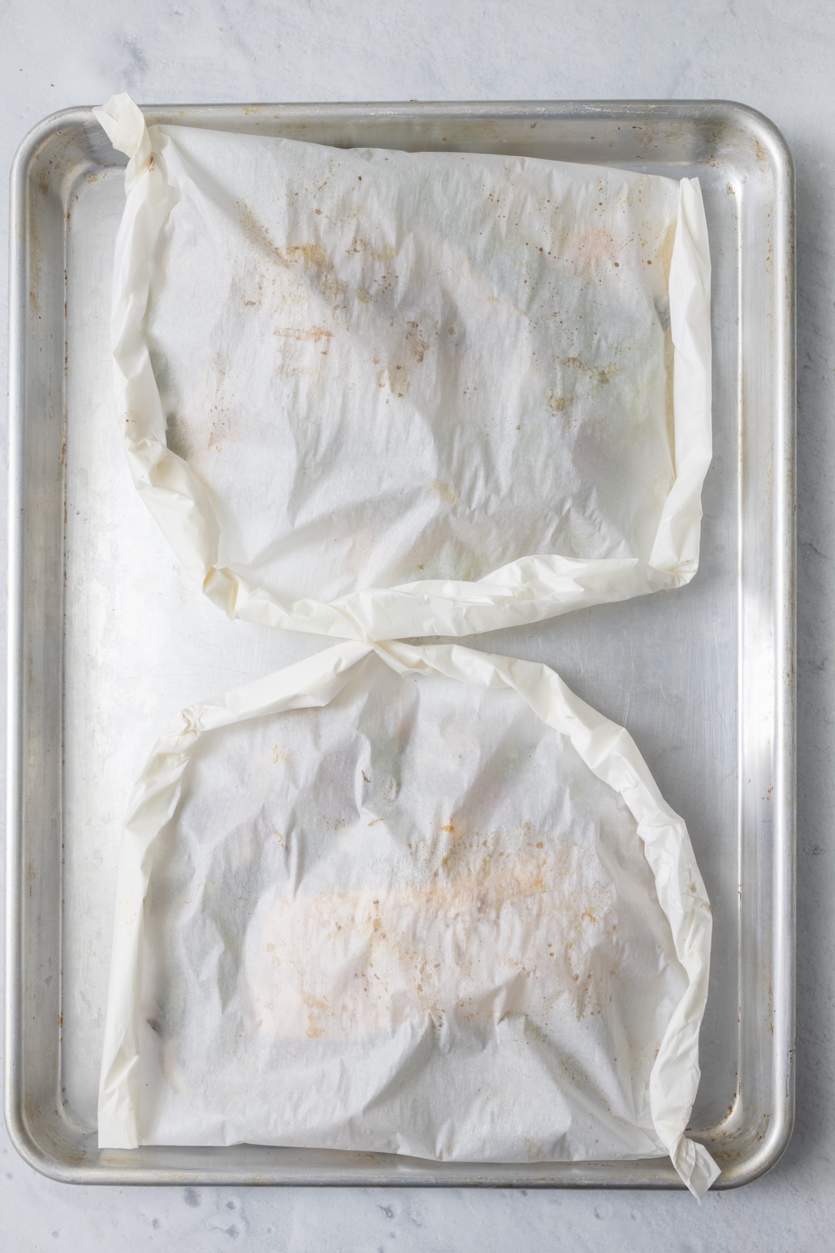 Two sealed parchment paper fish packets on an aluminum baking sheet ready for the oven.