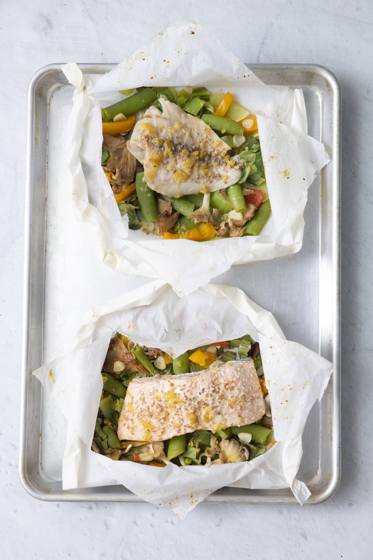 Two opened fish in parchment paper packets on a baking sheet showing cooked whitefish and salmon with vegetables after baking.