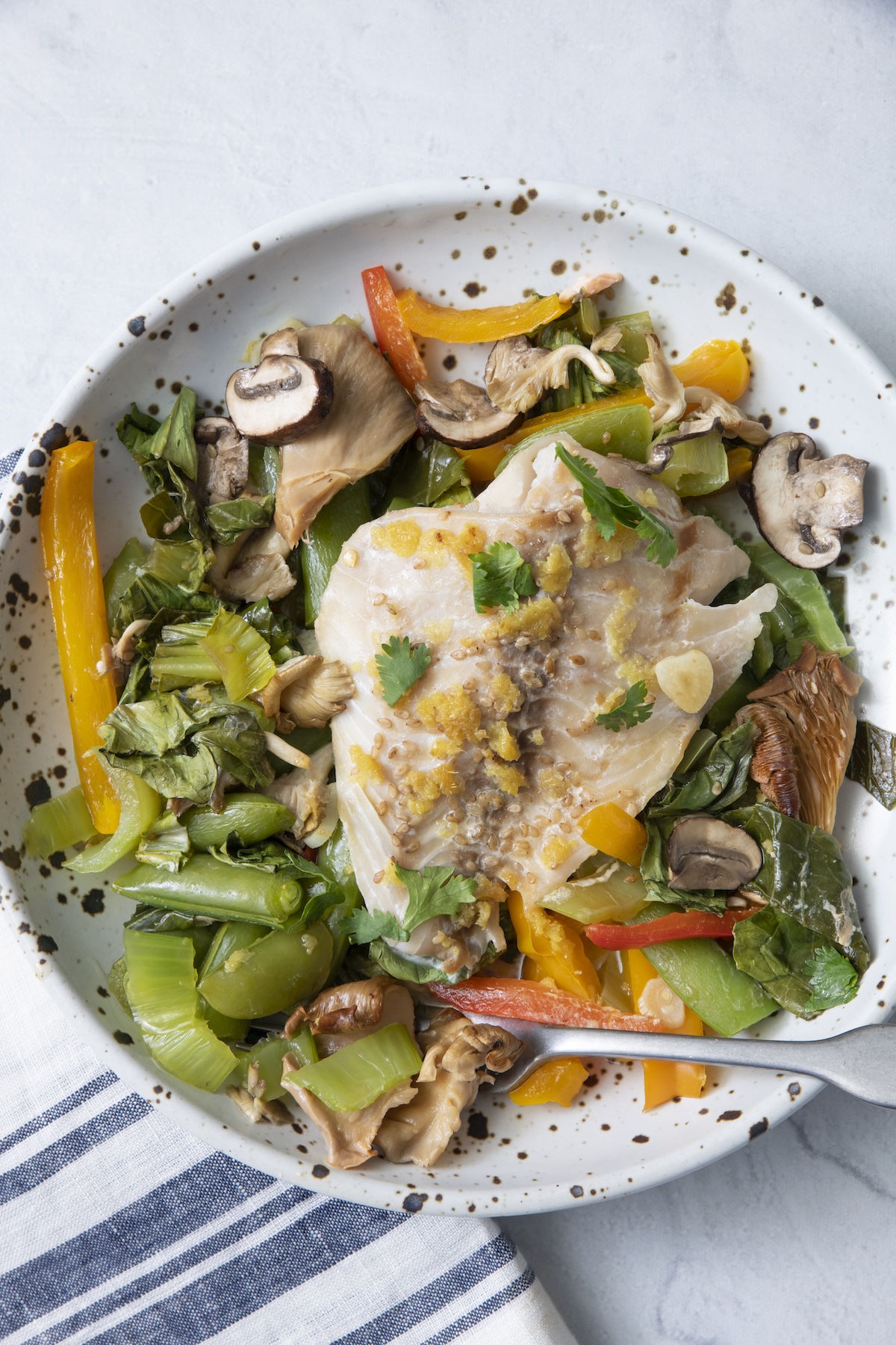 Fish in parchment paper recipe served in a white speckled bowl with bok choy, mushrooms, snap peas, and bell peppers on a marble surface.