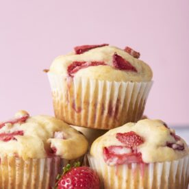 Three freshly baked strawberry muffins stacked with juicy strawberry pieces on top and a fresh strawberry in front on a white surface.