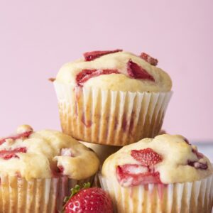 Three freshly baked strawberry muffins stacked with juicy strawberry pieces on top and a fresh strawberry in front on a white surface.