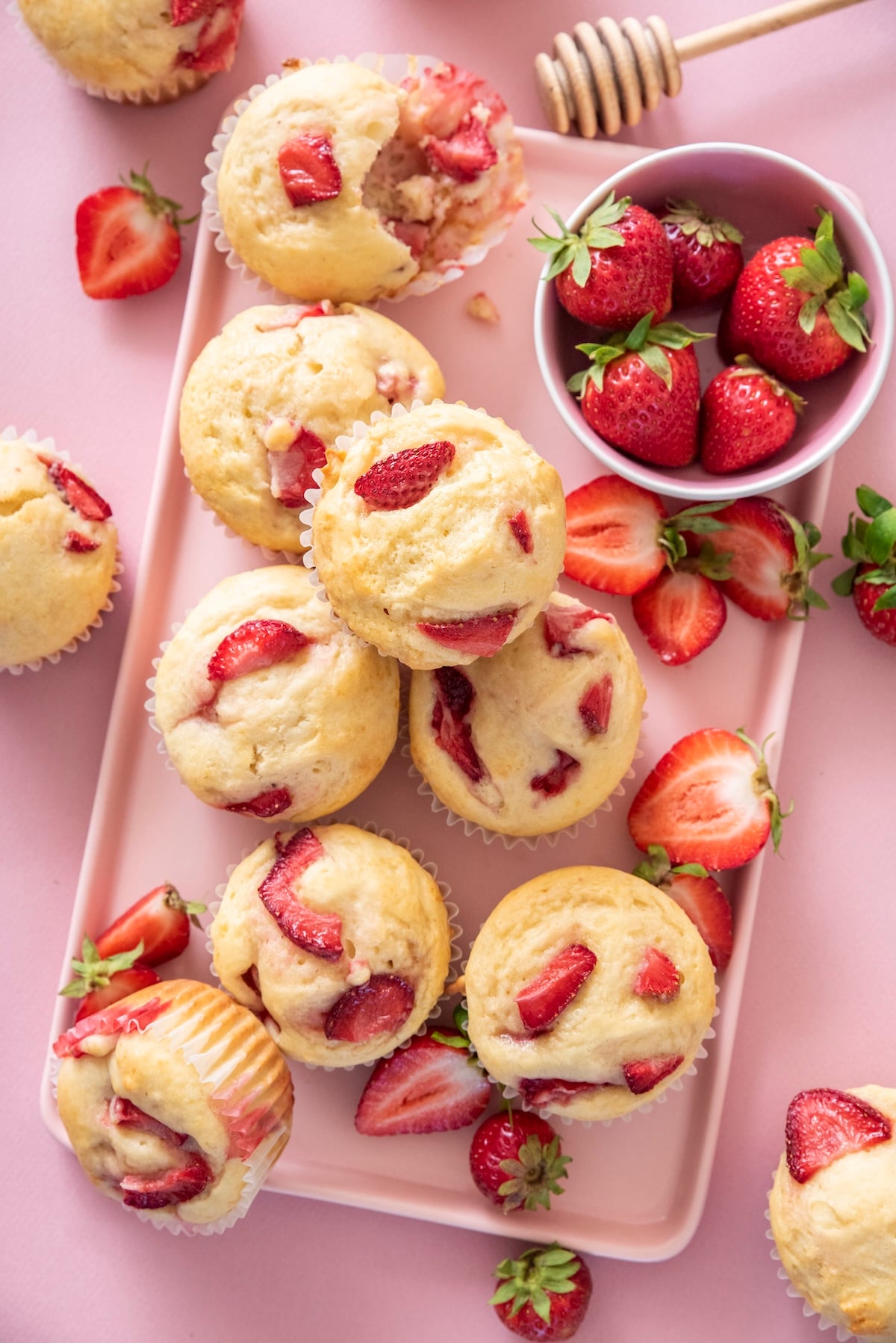 Overhead view of strawberry muffins on a pink tray surrounded by fresh strawberries and a honey dipper, one muffin with a bite taken out.