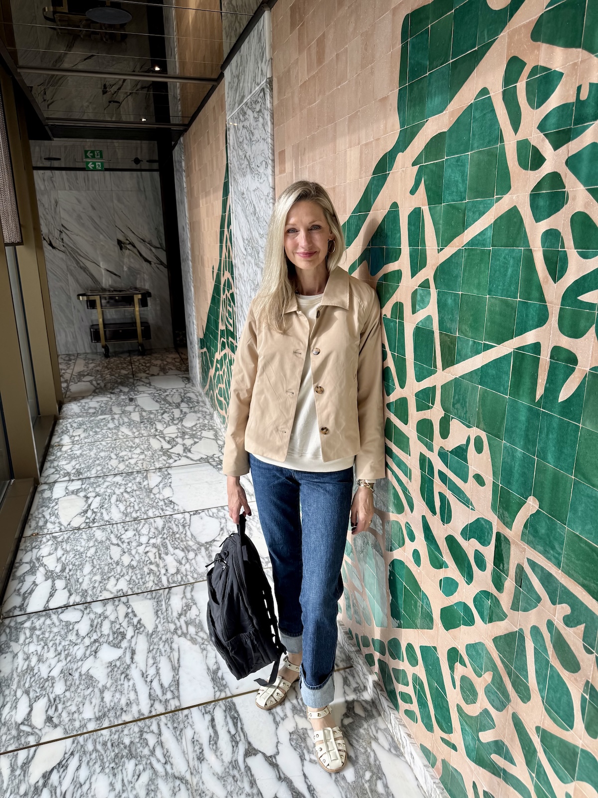 Catherine posing in a tan jacket and jeans against a dramatic green and blush Moroccan tile wall.