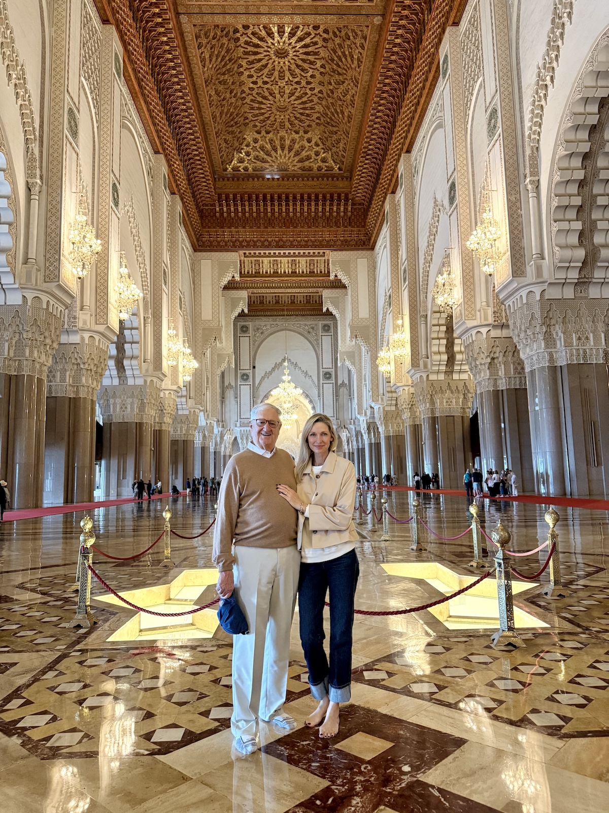 Catherine and her dad standing inside the Hassan II Mosque in Casablanca, the grand carved ceiling, marble floors, and golden chandeliers stretching behind them