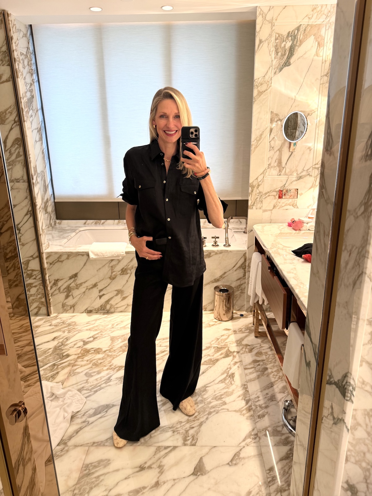 Catherine taking a mirror selfie in a black linen shirt and wide-leg trousers in a marble hotel bathroom.