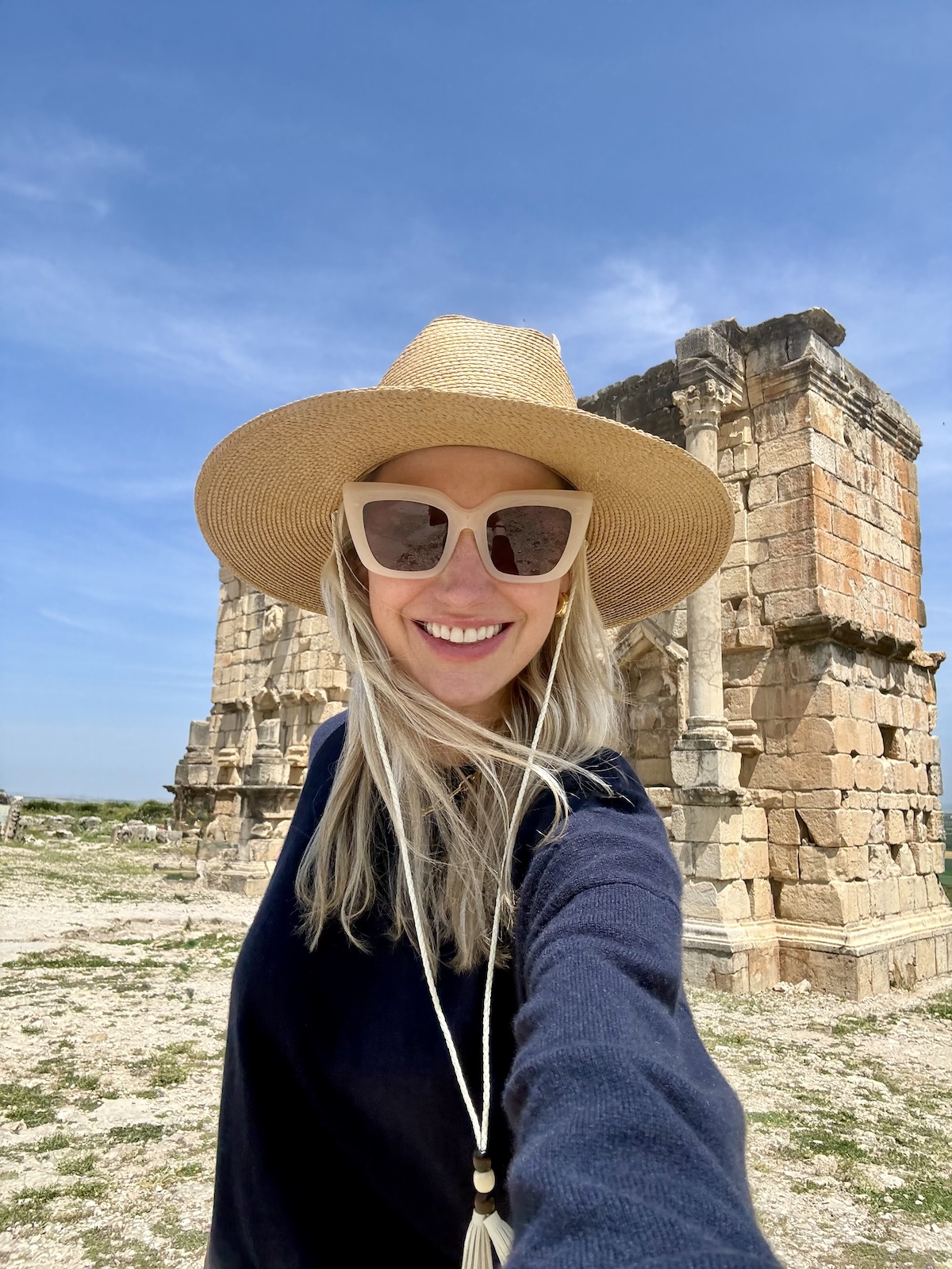 Catherine taking a selfie at the Roman ruins of Volubilis, wearing a straw hat and sunglasses, ancient stone columns behind her.