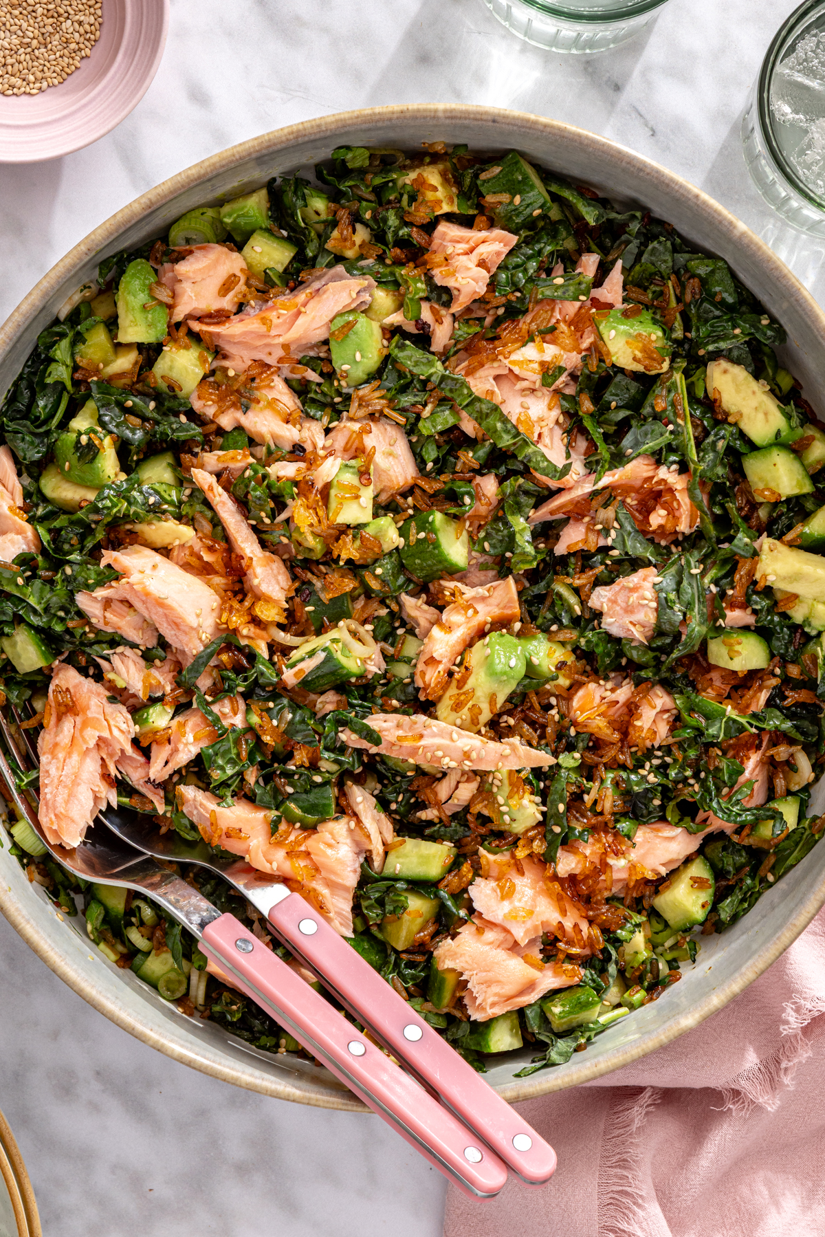 Crispy rice salmon salad with broiled salmon, kale, avocado, and cucumber in a large bowl with pink serving tongs.