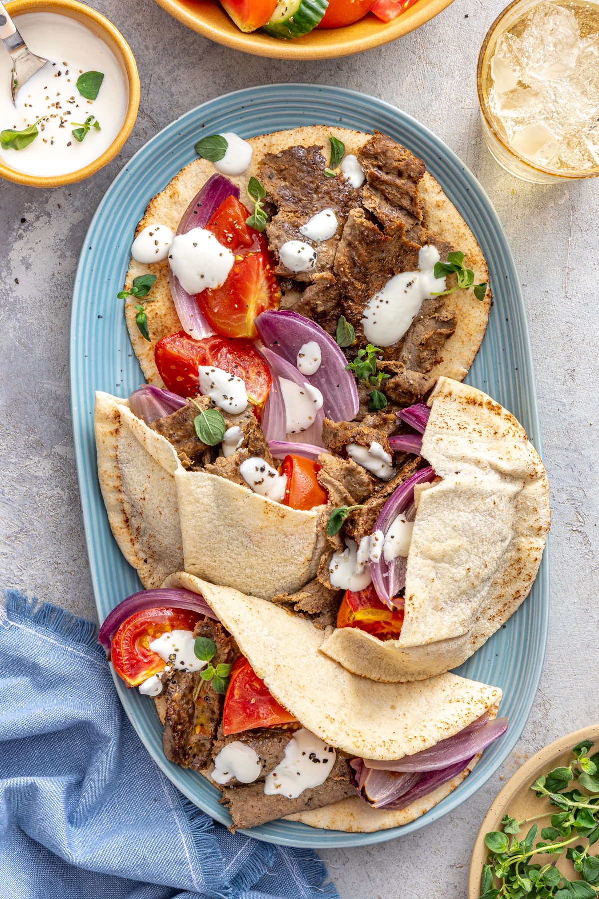 Homemade doner kebab served on warm pita bread with roasted tomatoes, red onion, and lemony yogurt sauce on a blue oval platter.