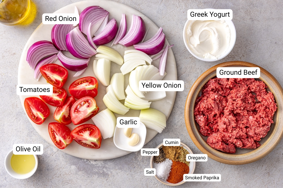 Homemade doner kebab ingredients including ground beef, yellow onion, red onion, tomatoes, garlic, Greek yogurt, olive oil, and spices arranged on a marble board.