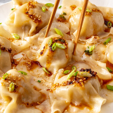 Chopsticks lifting a blanket dumpling from a plate topped with chili crisp and scallions.