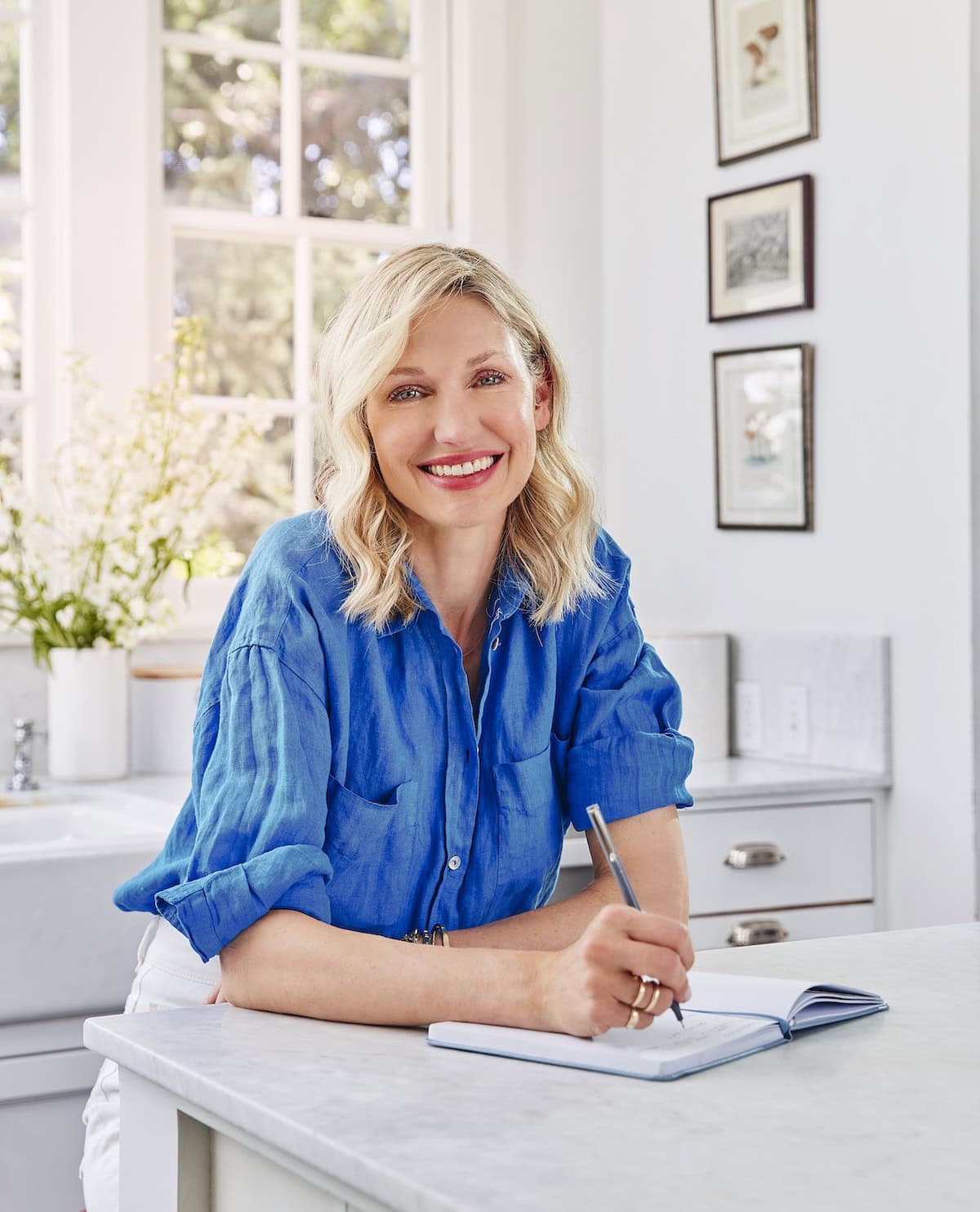 Catherine McCord in a bright blue shirt sits at a kitchen counter, writing in a notebook.