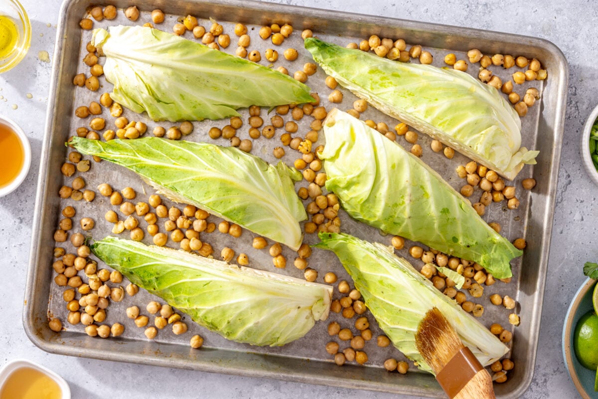 Six cabbage wedges and chickpeas on a baking sheet, about to be brushed with oil or sauce. Small bowls with ingredients surround the pan on a light countertop.