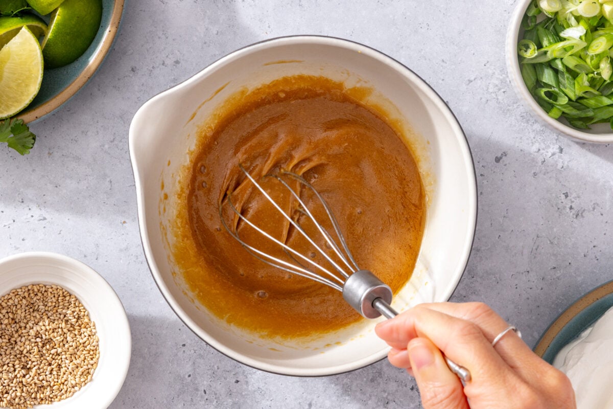A hand whisks a thick brown sauce in a white bowl. Around the bowl are ingredients including sliced green onions, sesame seeds, and lime wedges on a light-colored countertop.