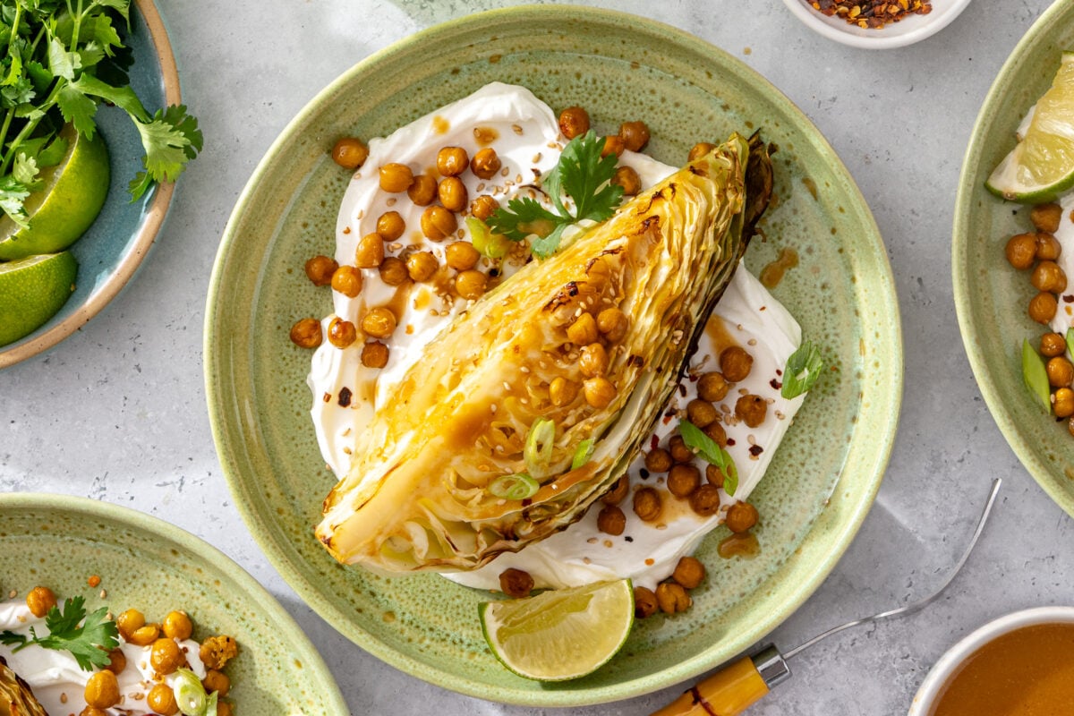A charred wedge of cabbage on a plate with creamy sauce, roasted chickpeas, fresh cilantro, green onions, red pepper flakes, and a lime wedge. Other plates and ingredients surround the main dish.